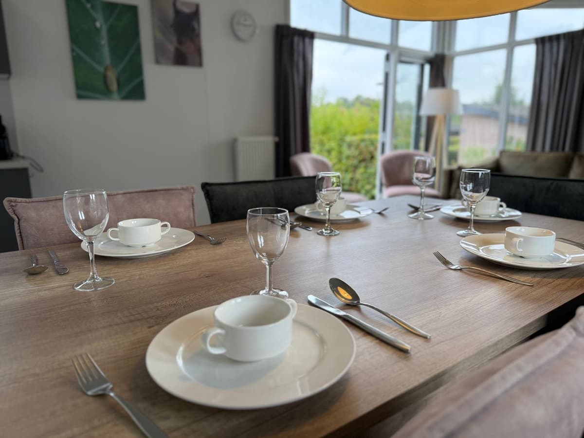 Eettafel met servies, glazen en bestek in een lichte woonkamer met grote ramen en groene omgeving.