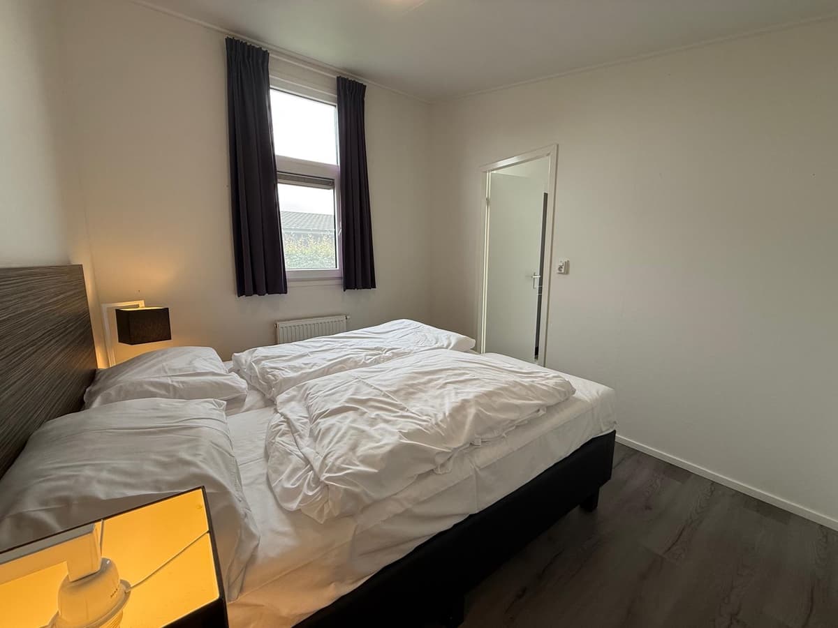 Slaapkamer met tweepersoonsbed, nachtkastje met lamp en raam met gordijnen aan de muur.