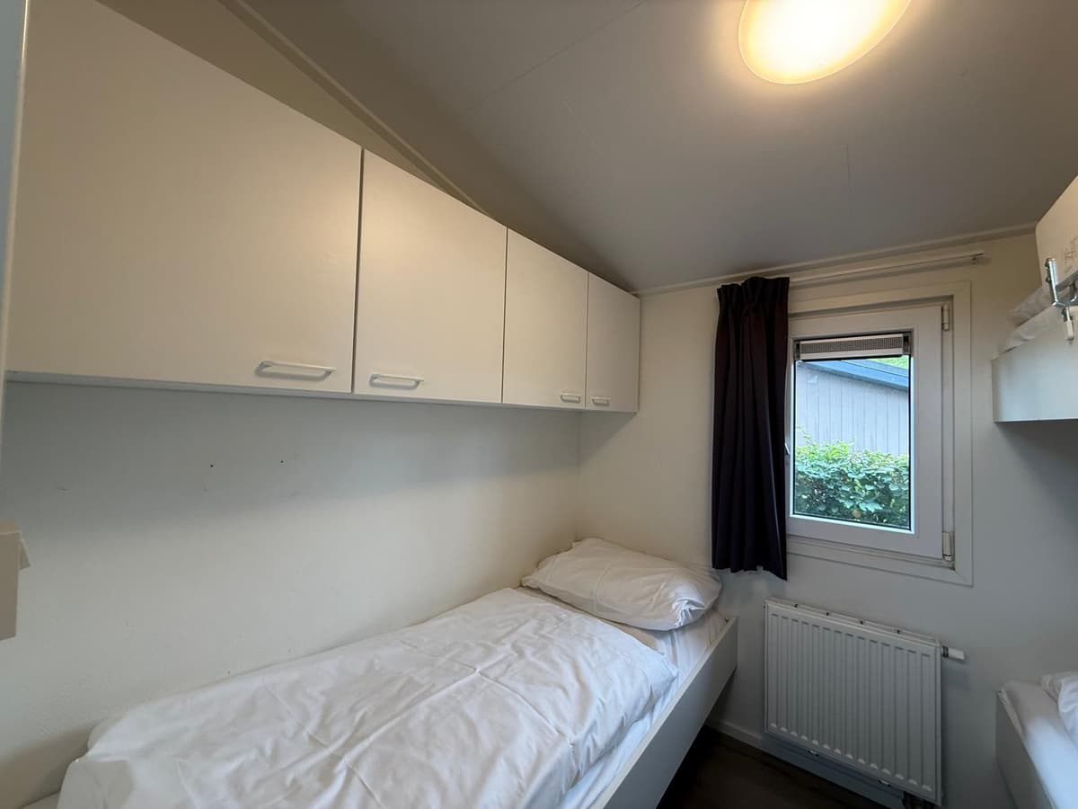 Slaapkamer met twee eenpersoonsbedden, eenvoudige witte kasten en een raam met uitzicht op groen buiten.