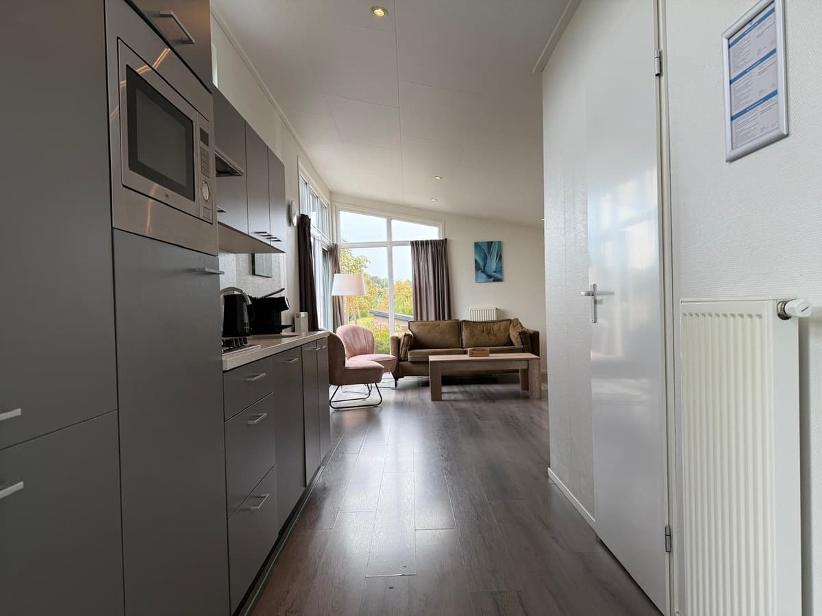 Ruime woonkamer met keuken, bank, fauteuil en grote ramen met uitzicht naar buiten.