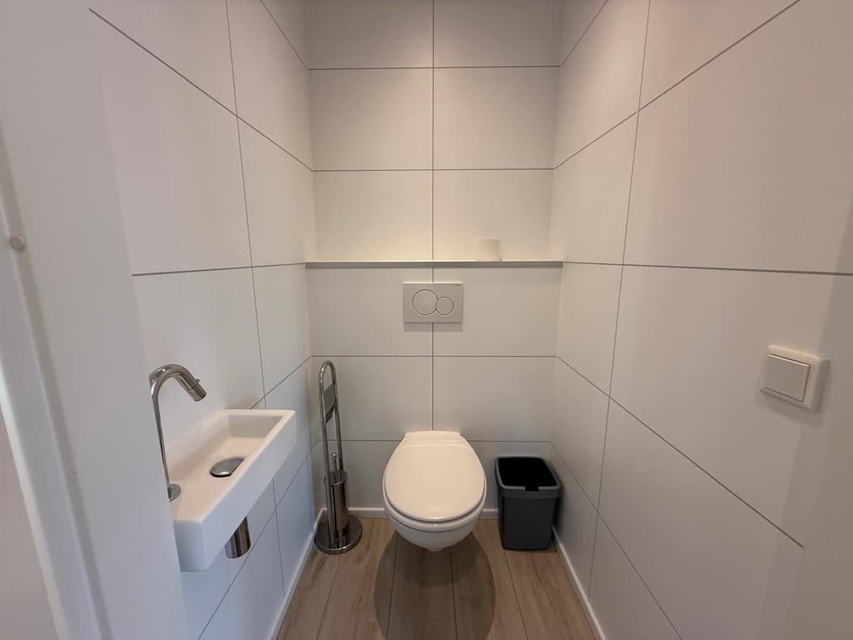 Moderne toiletruimte met witte tegels, een wandtoilet, een fonteintje en een prullenbak.