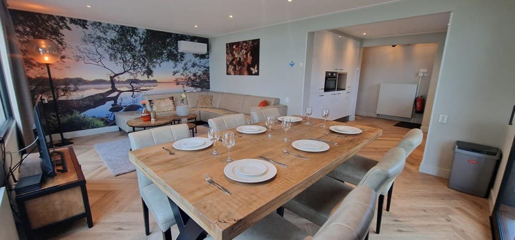 Gezellige eet- en woonkamer met grote eettafel, bank, TV en uitzicht op een natuurwanddecoratie.