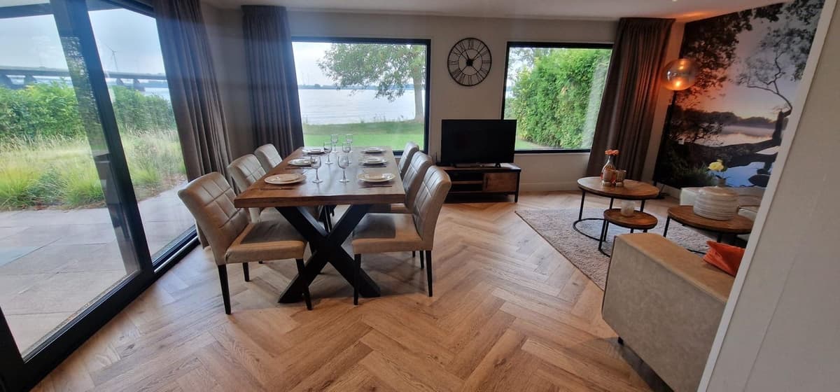 Eetkamertafel met stoelen, grote ramen met uitzicht op water en groene omgeving, televisie en moderne inrichting.