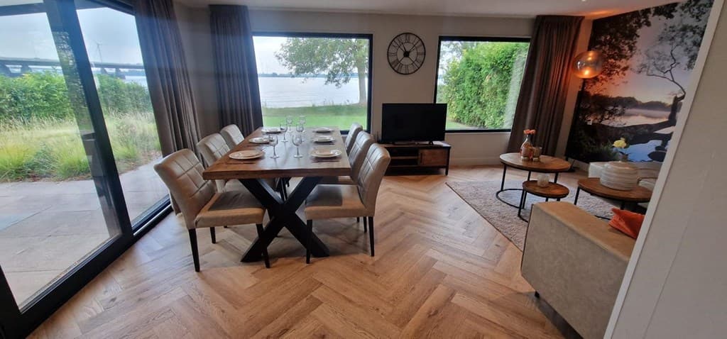 Eetkamertafel met stoelen, grote ramen met uitzicht op water en groene omgeving, televisie en moderne inrichting.