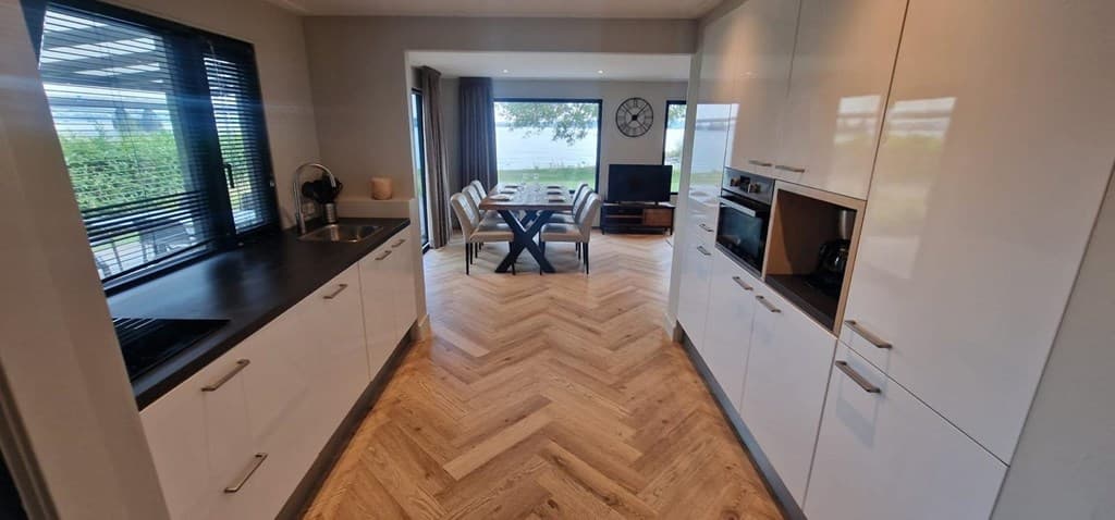 Moderne keuken met wit keukenmeubilair, houten vloer en uitzicht op een eetgedeelte met grote ramen en een televisie.