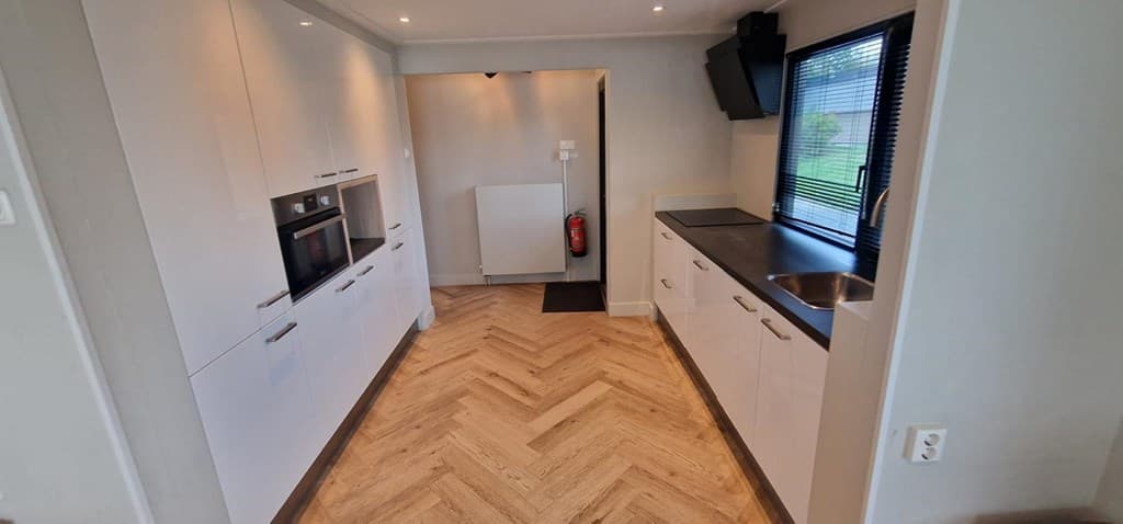 Moderne keuken met witte kasten, licht houten vloer en zwart aanrechtblad, voorzien van oven en raam met uitzicht op buiten.