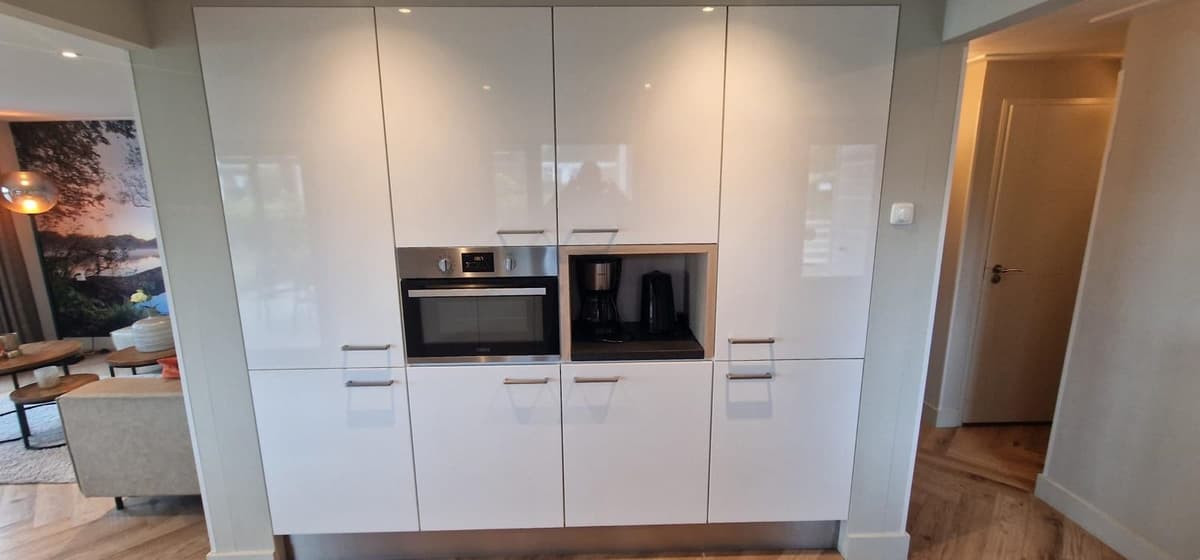 Moderne keukenwand met inbouwapparatuur zoals oven, magnetron en koffiezetter, omgeven door strakke, witte kasten