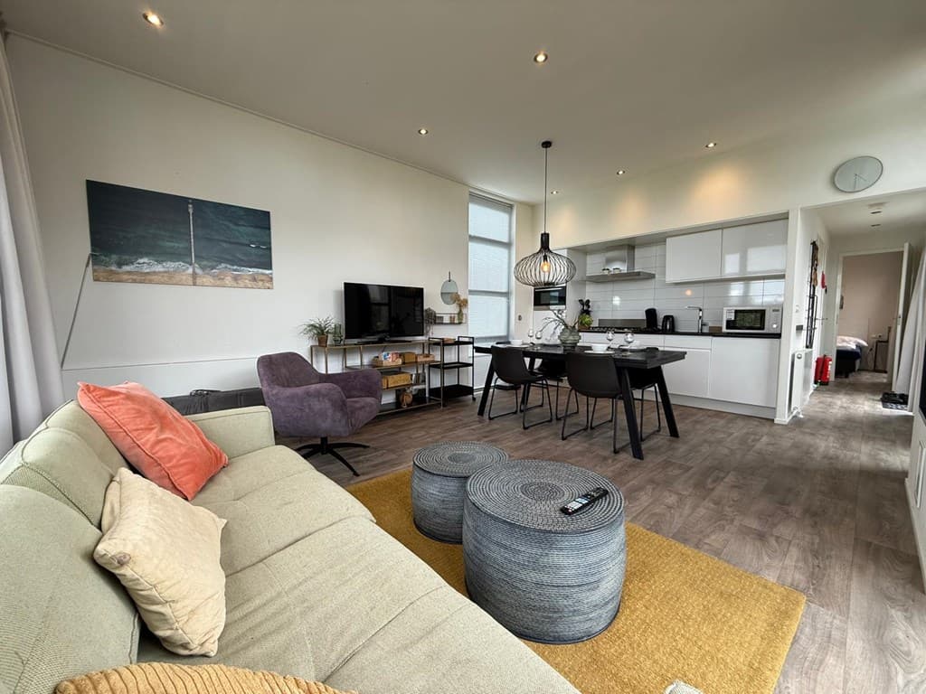 Moderne woonkamer met comfortabele bank, televisie, eettafel, en keuken; veel licht door grote ramen.