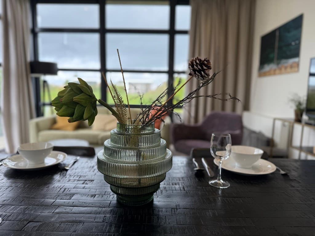 Eettafel met decoratieve vaas en takken, uitzicht op een ruime woonkamer met bank en grote ramen