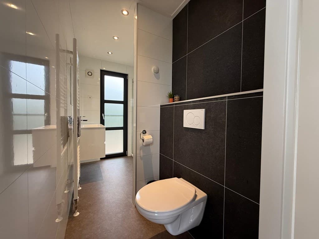 Moderne badkamer met betegelde wanden, toilet en toegang tot een lichte ruimte met een raam.
