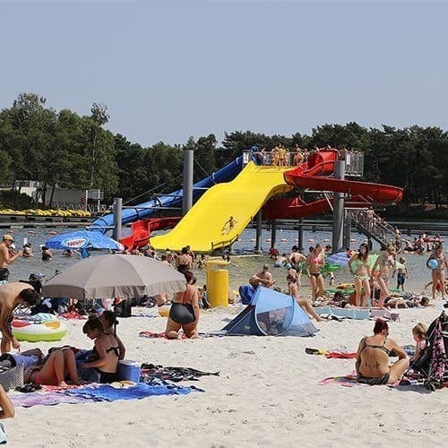 Strand met veel liggende en spelende mensen, op de achtergrond een waterglijbaan en een helderblauwe zwemvijver
