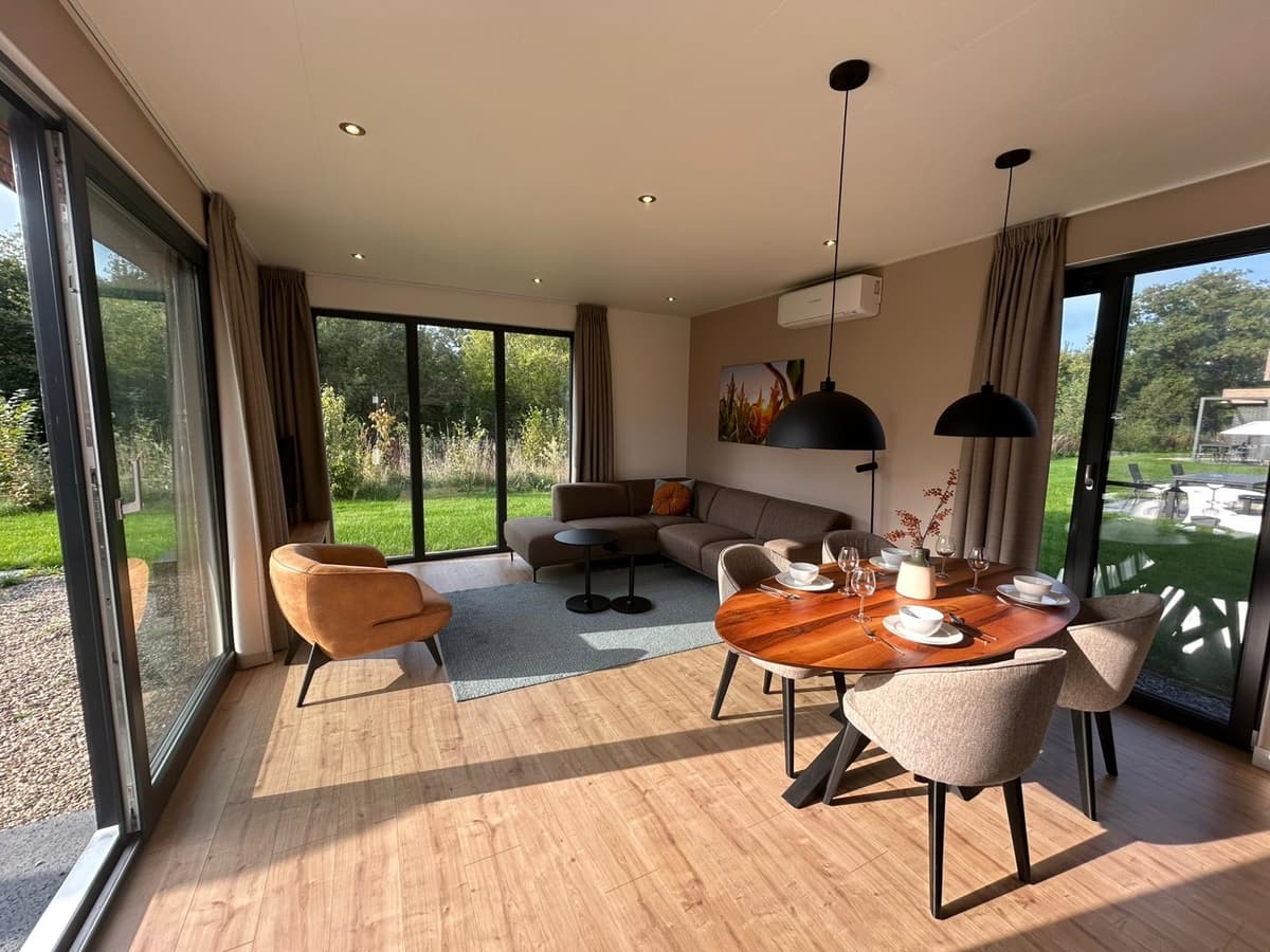 Moderne woonkamer met grote ramen, bank, eetgedeelte en uitzicht op groene omgeving in vakantiepark Hofparken Wiltershaar.