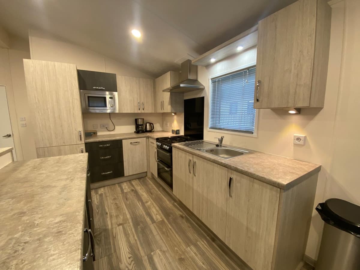 Moderne keuken met houten kasten, kookplaat, oven, magnetron en spoelbak met raam en verlichting boven het aanrecht.