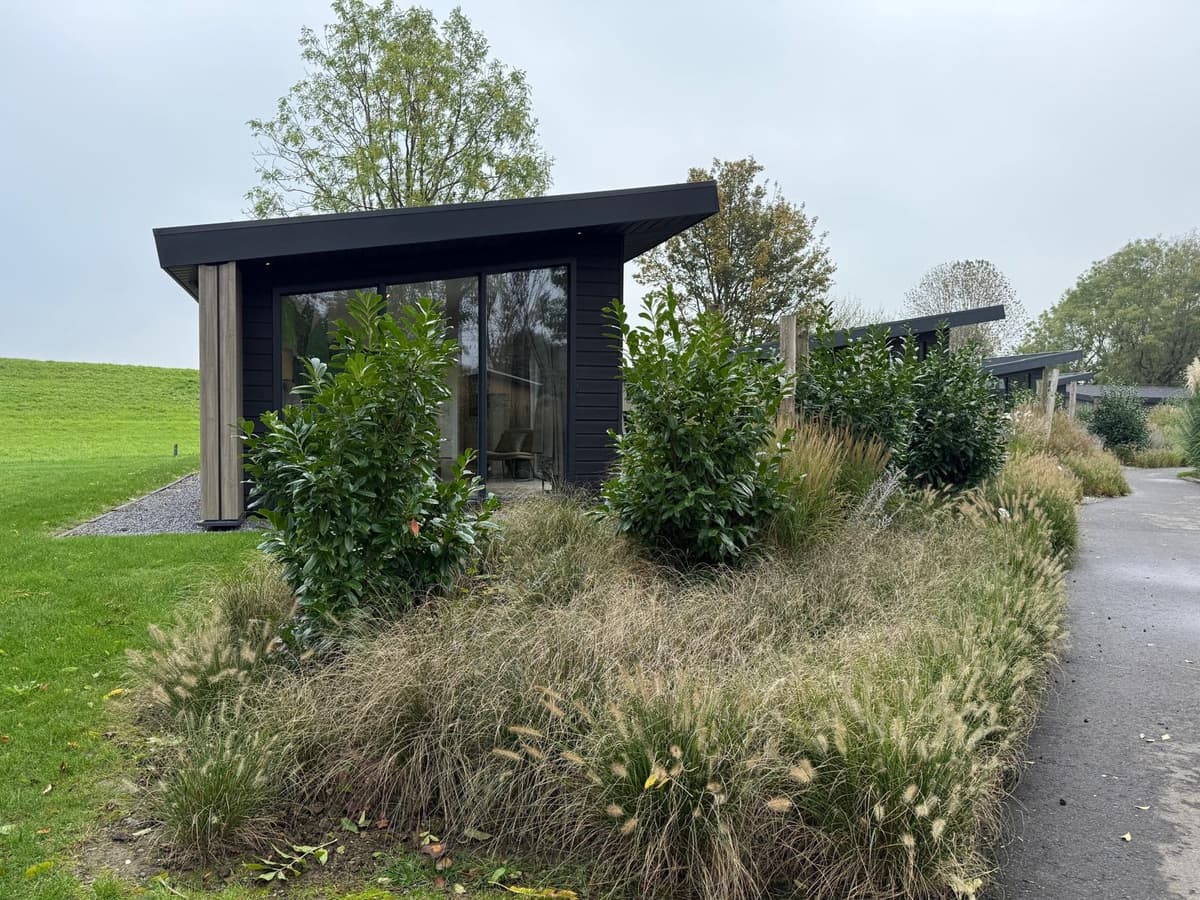 Moderne vakantiewoningen met grote ramen, omgeven door groen en struiken, gelegen aan een rustige laan.