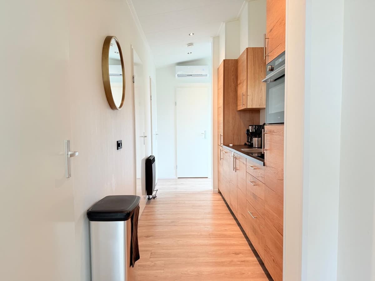 Lichte gang met een houten vloer, moderne keukenunits en een ronde spiegel aan de muur.