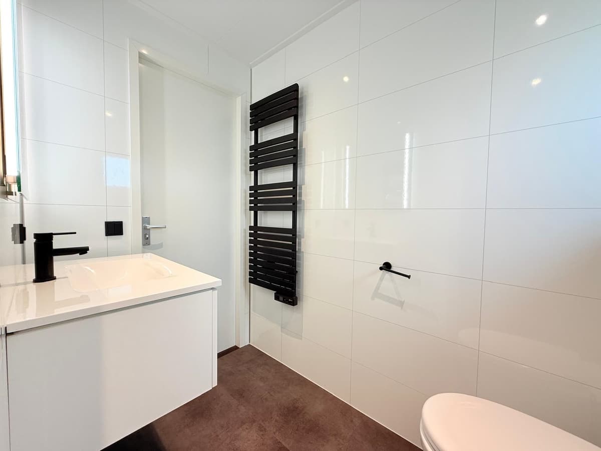Moderne badkamer met witte tegels, wastafel, zwarte kranen en handdoekradiator tegen de muur.