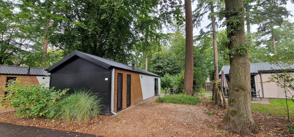 Huis met houten accenten en donkere gevel, omringd door bomen en een grindpad met groen.