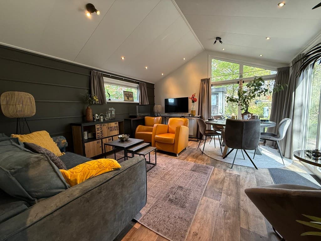 Gezellige woonkamer met gele stoelen, een tv, een eethoek en veel natuurlijke lichtinval door grote ramen.