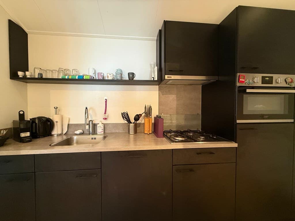 Moderne keuken met zwarte kastjes, rvs spoelbak, kookplaat, oven en een schap met glazen en keukenaccessoires.