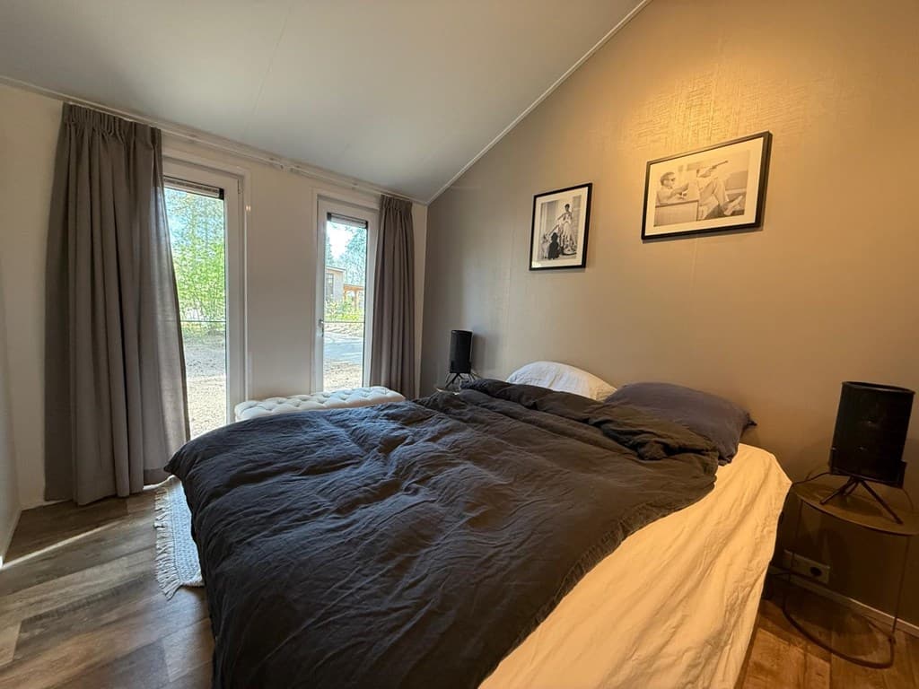 Slaapkamer met een tweepersoonsbed, natuurlicht via twee ramen en houten vloer, voorzien van gordijnen en wanddecoratie.