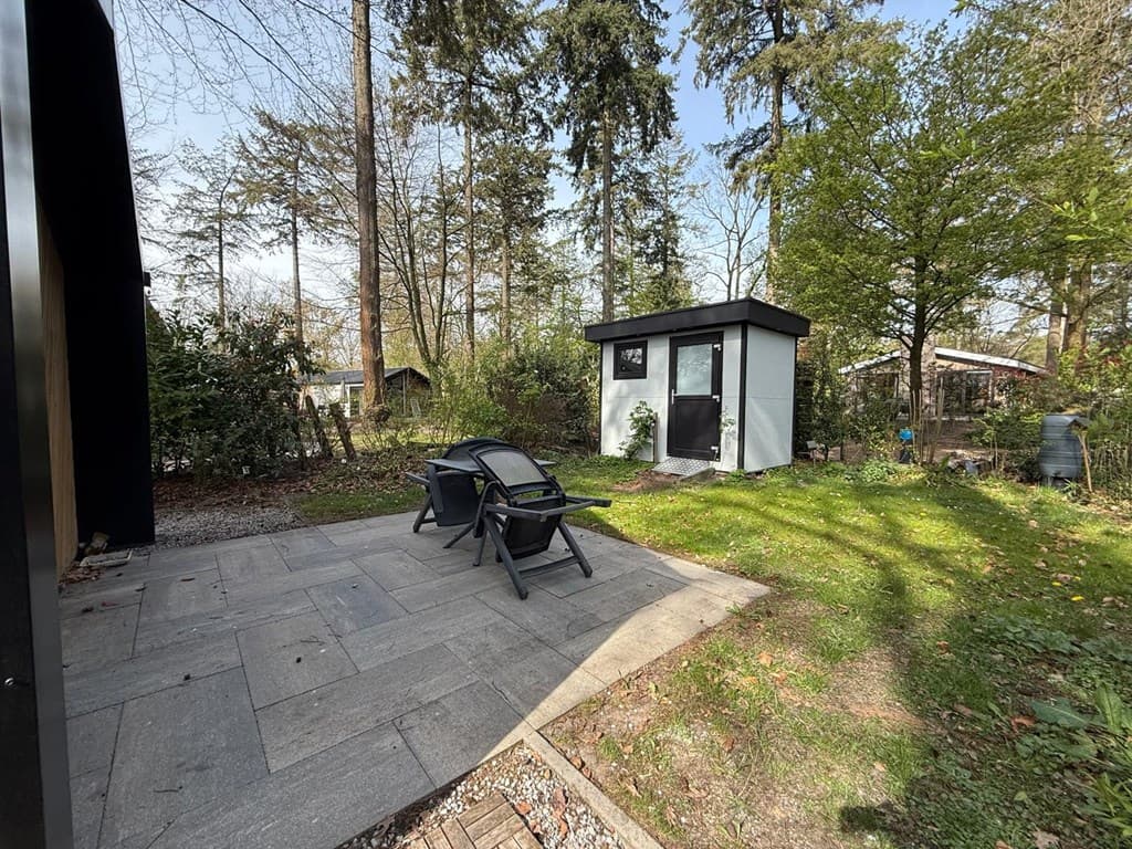 Tuin met grasveld, betegeld terras en tuinhuisje omgeven door bomen en beplanting
