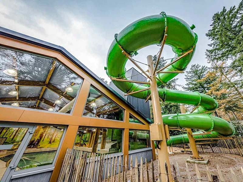 Glijbaan in een groene kleur, bevestigd aan een modern gebouw met veel glas en houten accenten, omgeven door bomen.