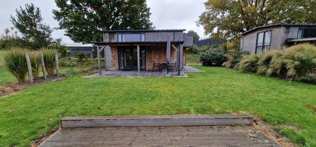 Vrijstaand vakantiehuis met houten gevel en grote ramen, omgeven door een groene tuin met gras en struiken.