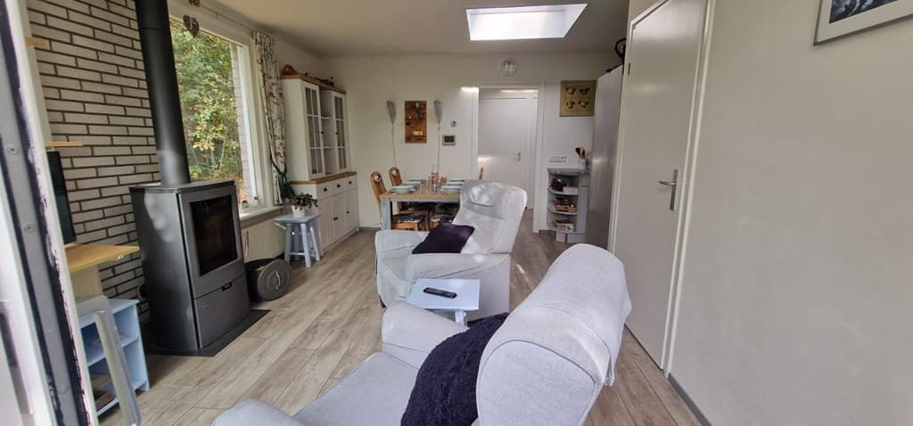 Woonkamer met banken, een houtkachel en een eettafel bij een raam met uitzicht op de natuur.