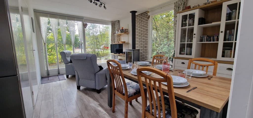 Eetkamer met houten tafel en zes stoelen, openhaard en uitzicht op de tuin door grote ramen.