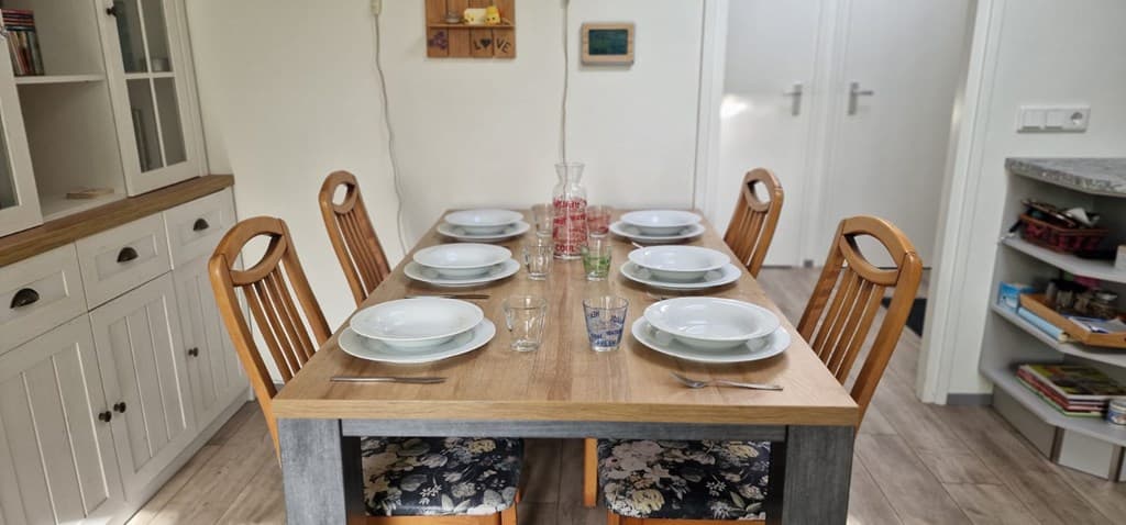 Eettafel met zes borden en glazen, omringd door houten stoelen in een lichte ruimte met een open keuken.