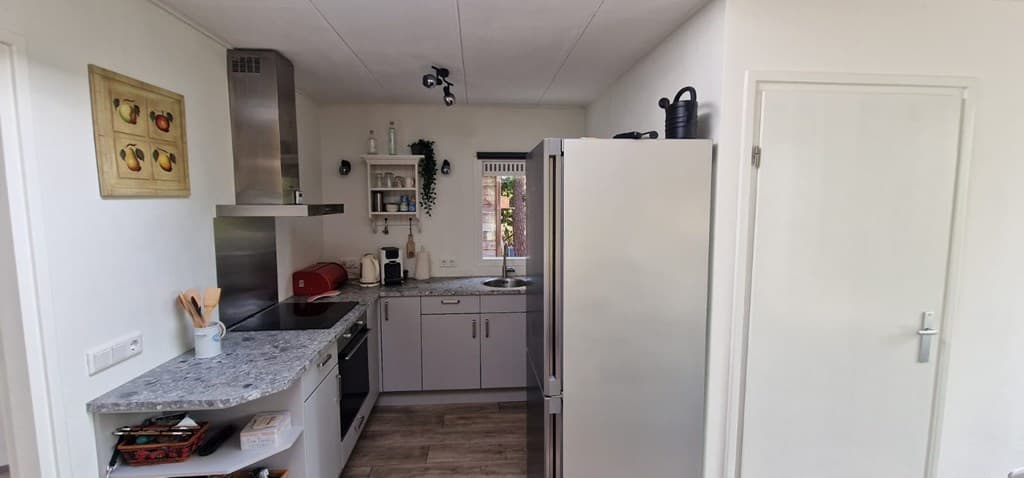 Moderne keuken met grijze kasten, een granieten werkblad en een grote koelkast, goed verlicht door natuurlijk licht.