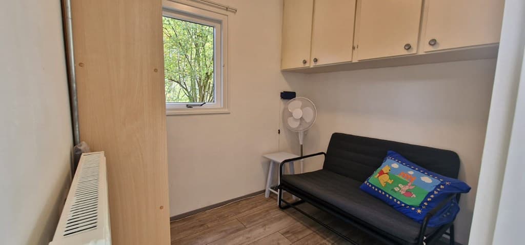 Kleine kamer met een bank, ventilator en kast, met een raam dat uitzicht biedt op bomen buiten