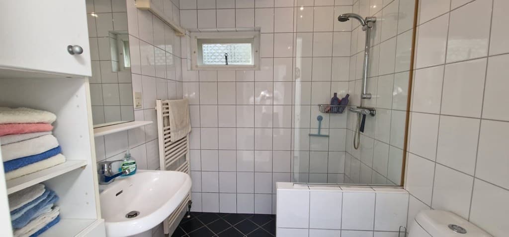 Badkamer met witte tegels, een wastafel, douchecabine, toilet en rek met handdoeken en toiletartikelen