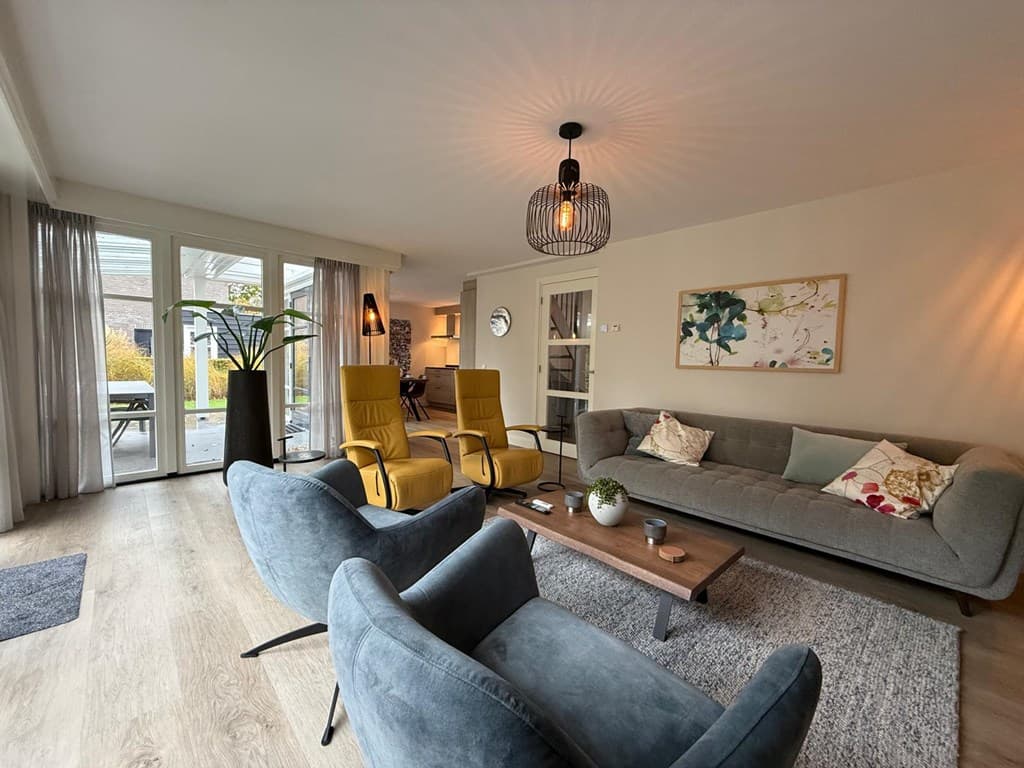 Gezellige woonkamer met grijze en gele fauteuils, een comfortabele bank, houten tafel en toegang tot een terras met uitzicht