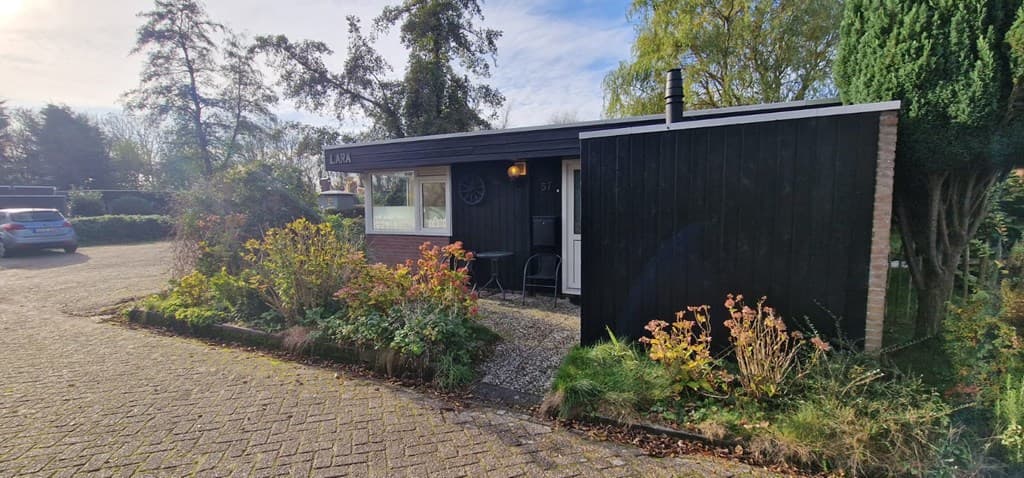 Zwart houten vakantiehuis met stenen details en een tuin, gelegen aan een grindpad met parkeergelegenheid nabij bomen.