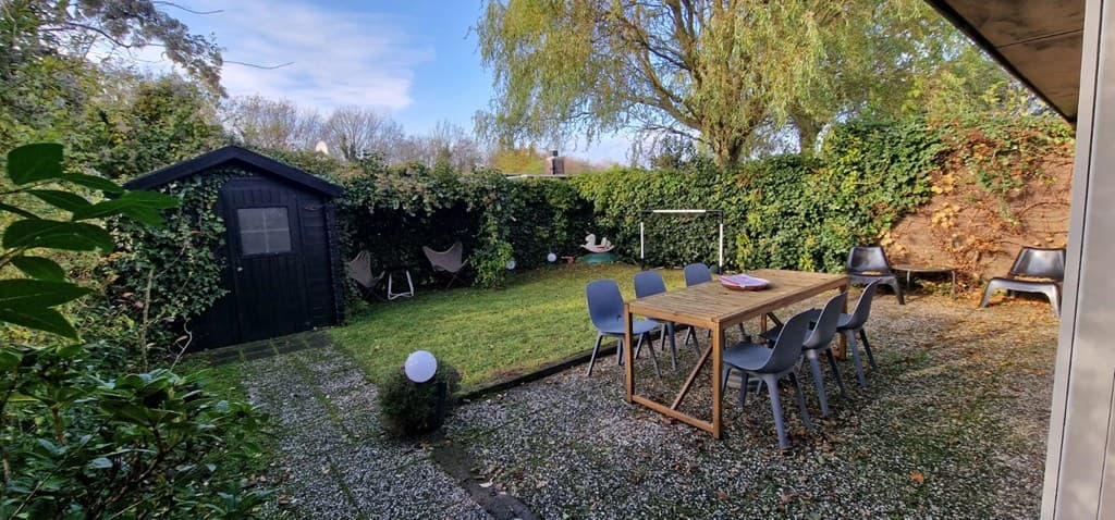 Tuin met gras en bestrating, een houten schuur, een grote tafel, stoelen en een speeltoestel.