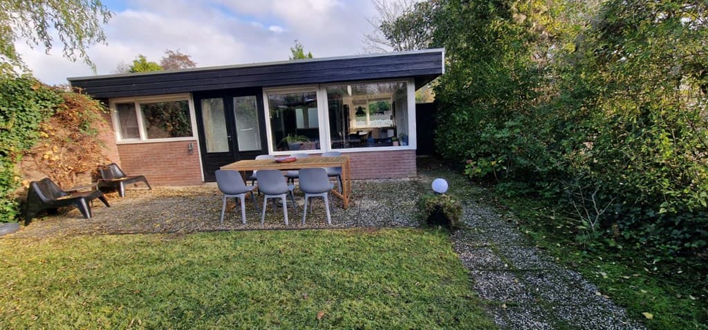 Aanzicht van een vakantiehuis met een terras en tuin, omringd door groen, met een tafel en stoelen.
