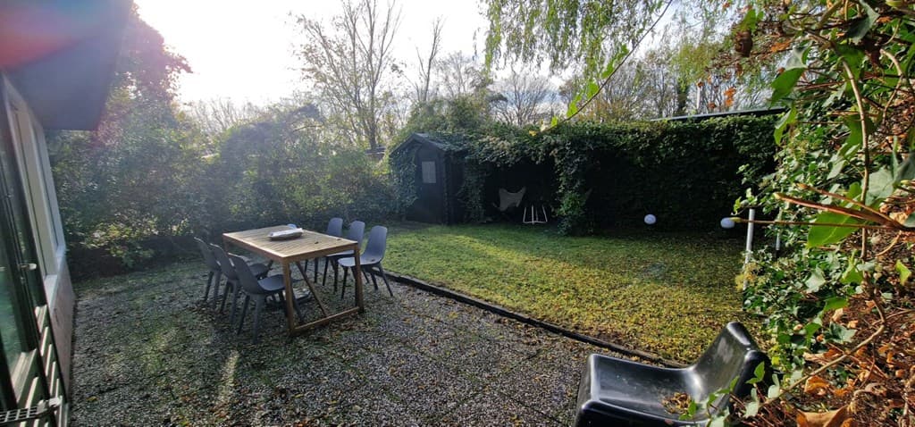 Tuin met gras en betegeld terras, een houten tafel met stoelen en een schuur omringd door begroeiing