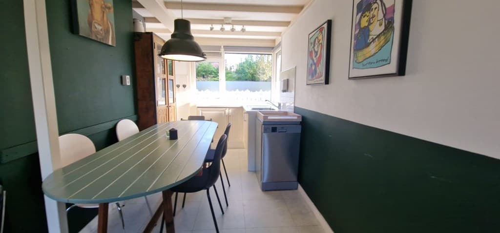 Eetruimte met lange tafel, stoelen, keuken met moderne apparatuur en kunst aan de wand.