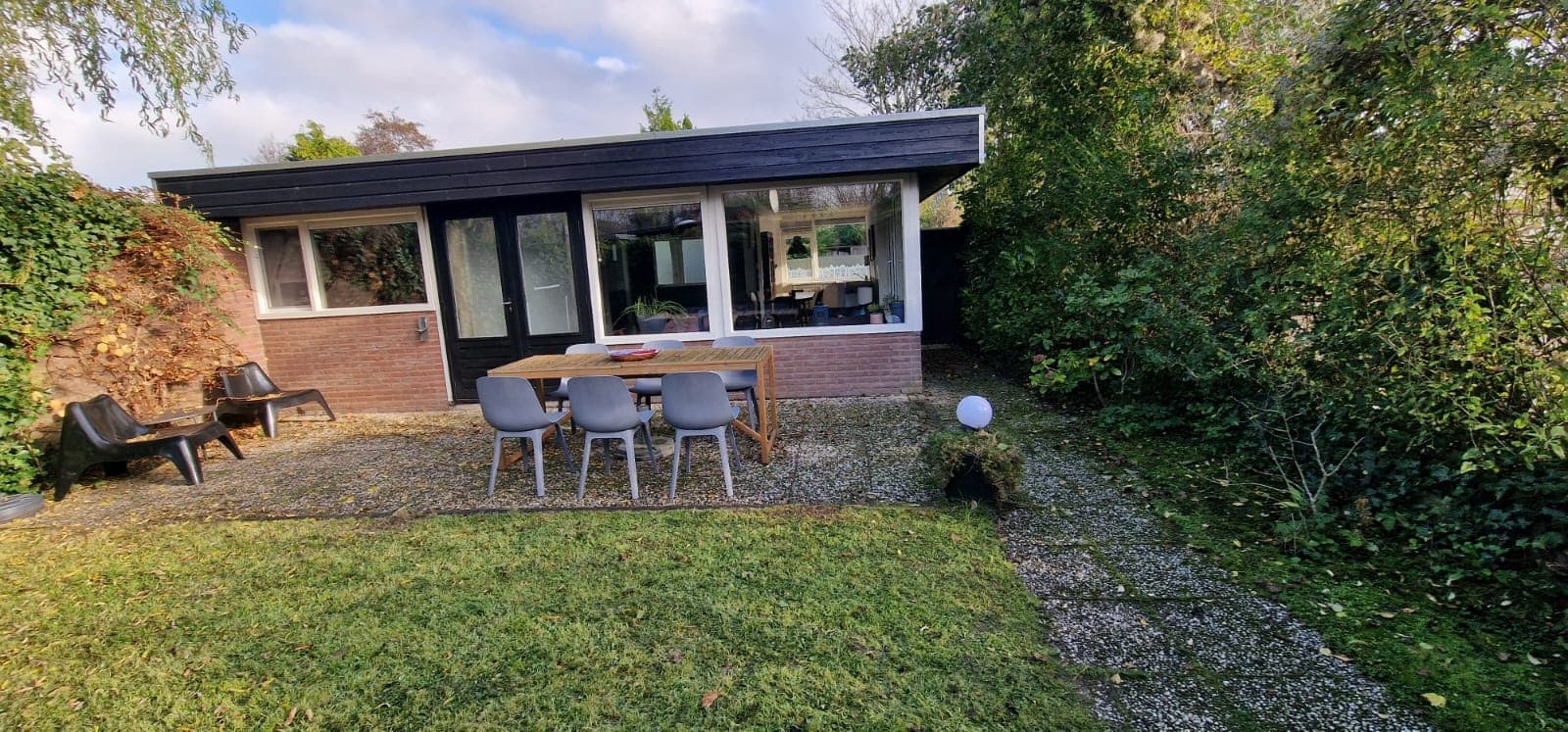 Aanzicht van een vakantiehuis met een terras en tuin, omringd door groen, met een tafel en stoelen.