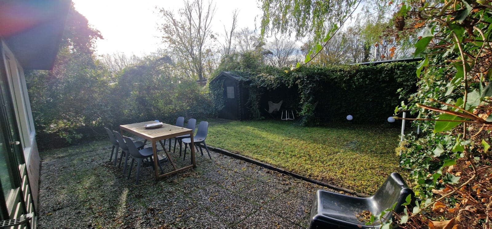 Tuin met gras en betegeld terras, een houten tafel met stoelen en een schuur omringd door begroeiing