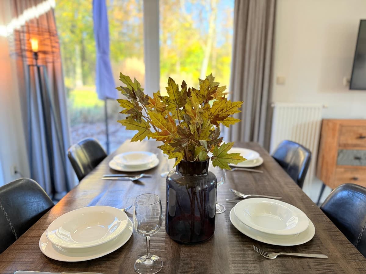 Eettafel met borden en glazen, een vaas met gele bladeren staat in het midden en grote ramen geven uitzicht op een groene omg