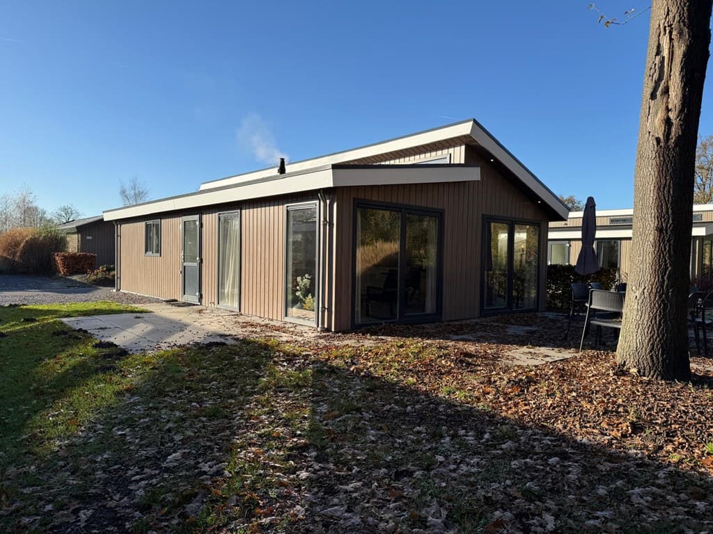 Moderne vakantiewoning met grote ramen, een terras en een tuin, omgeven door bomen en bladeren in het gras.