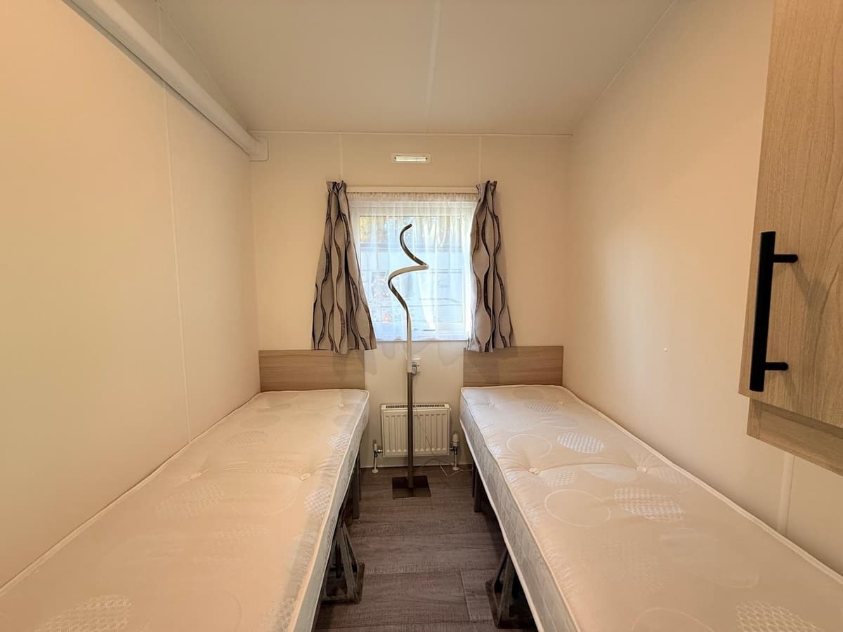 Slaapkamer met twee eenpersoonsbedden, een raam en een gordijn in lichte kleuren.