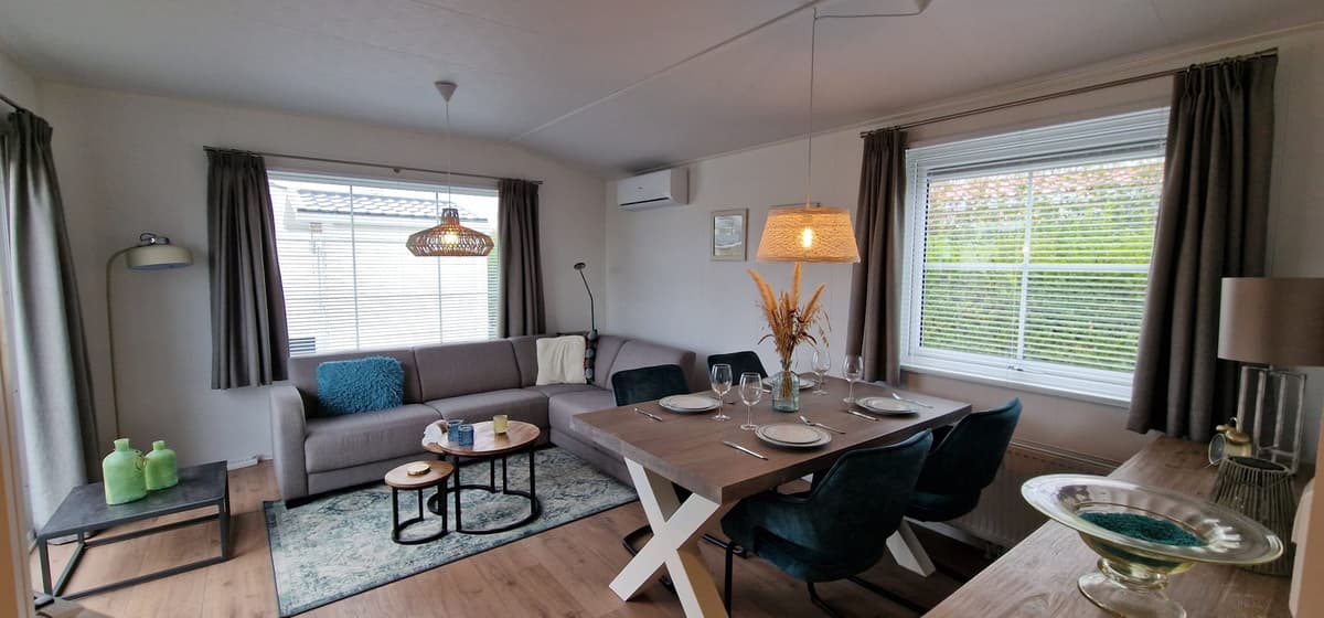 Woonkamer met een zithoek, eettafel en moderne decoraties, veel natuurlijk licht door de ramen.