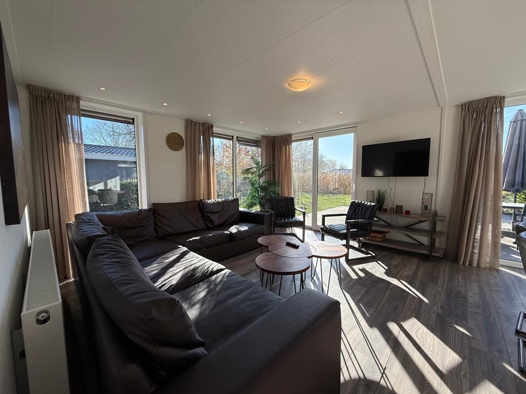 Zitkamer met een grote hoekbank, eettafel en tv, omgeven door ramen met veel lichtinval en uitzicht op de tuin