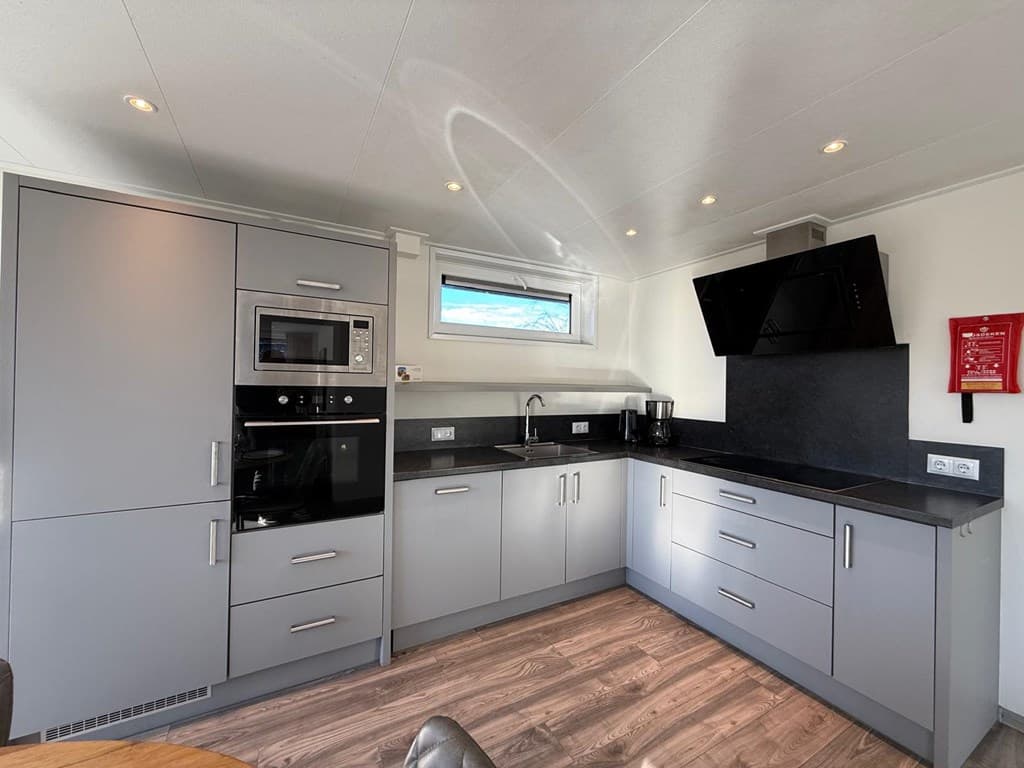 Moderne keuken met grijze kasten, een combimagnetron, oven, en zwarte werkbladen tegen een lichte wand.