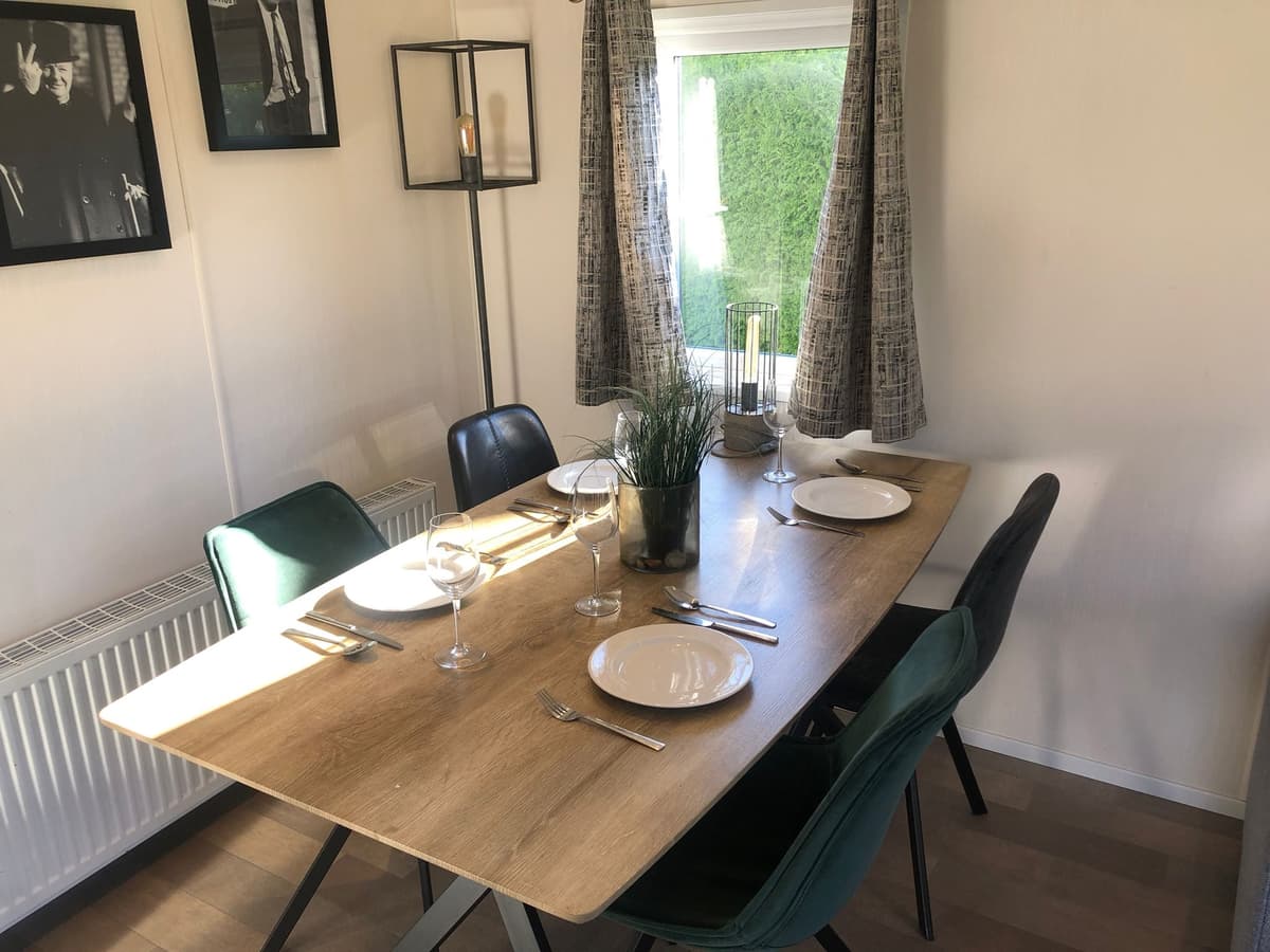 Eetkamertafel met glazen, borden en stoelen, raam met gordijnen, omringd door foto's aan de wand.