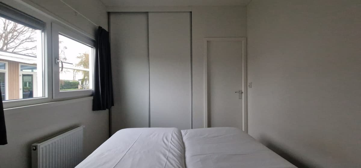 Slaapkamer met twee eenpersoonsbedden, witte muren en een inbouwkast, lichtinval via ramen aan de zijkant.