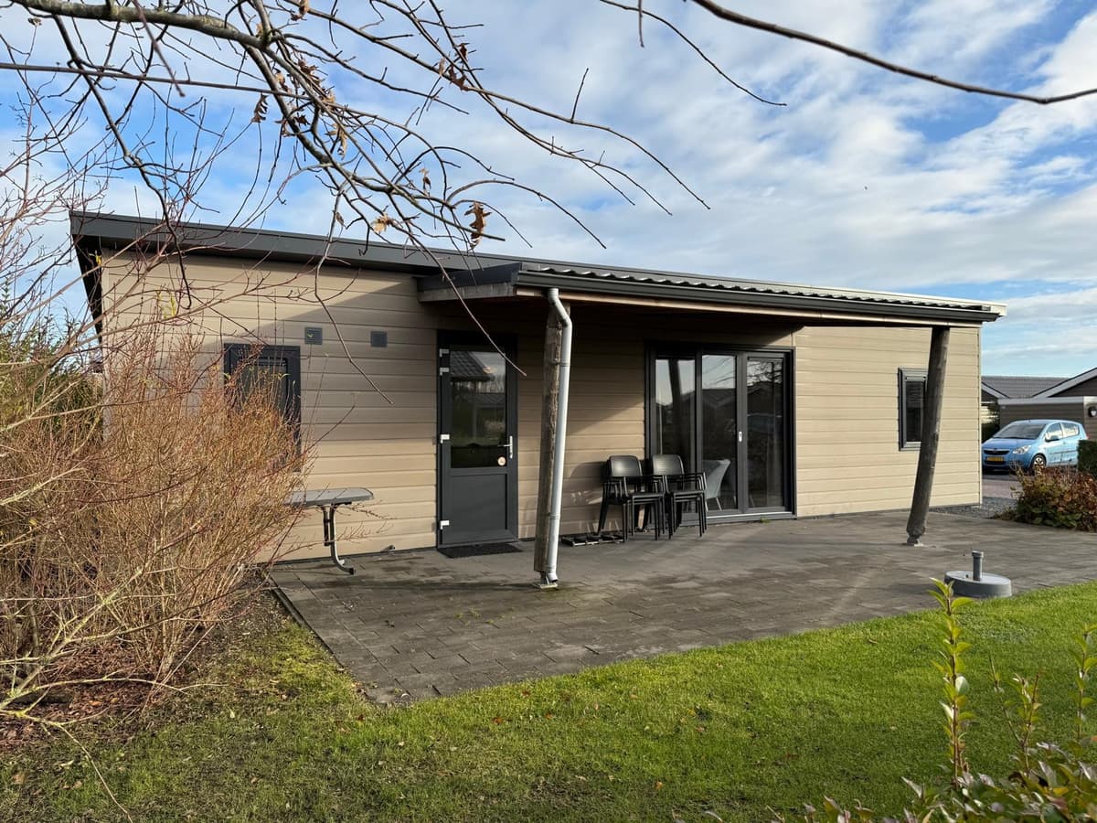 Moderne vakantiewoning met ruime veranda, tafel en stoelen, omgeven door gras en struiken.